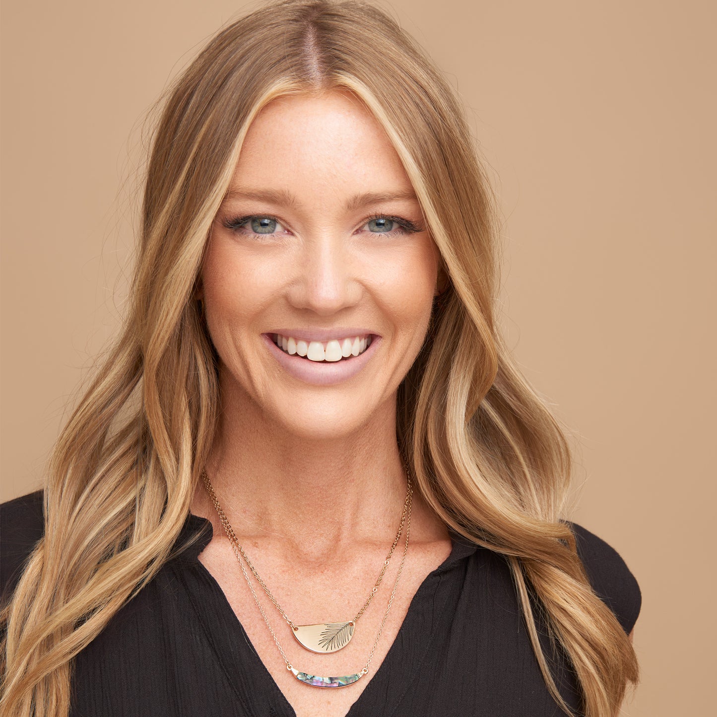 Woman with long blonde hair wearing a black top and layered necklaces on a beige background
