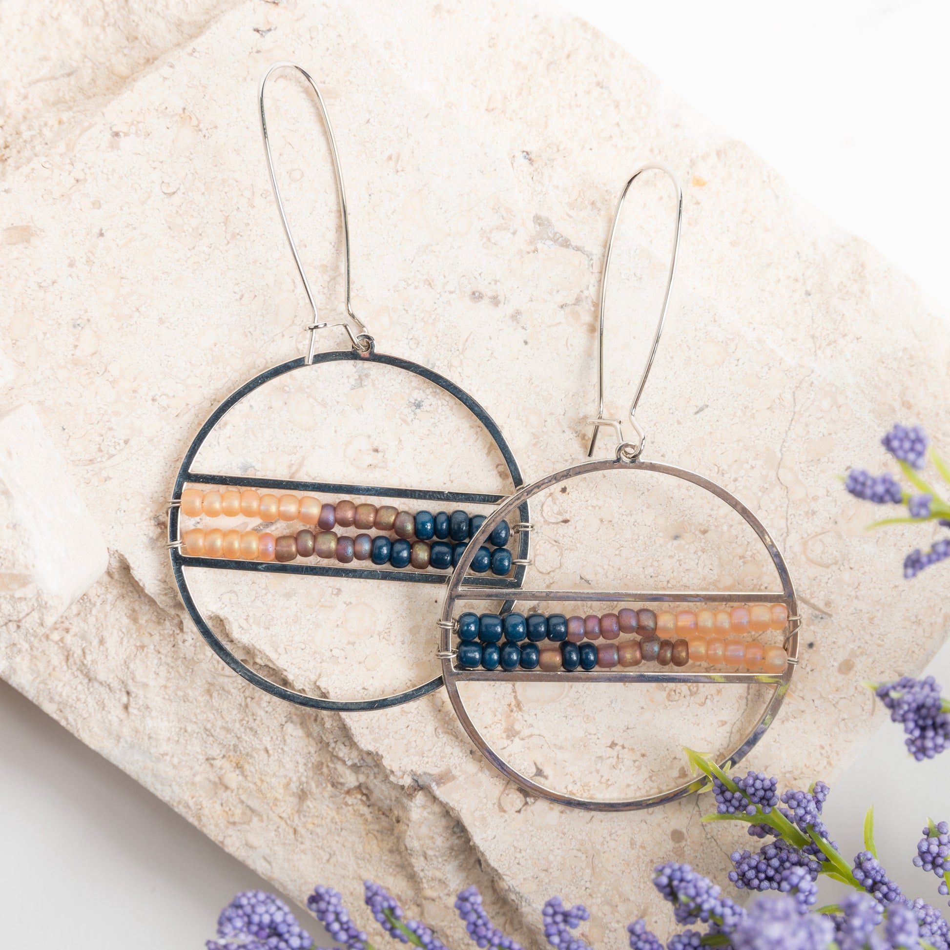 A pair of hoop earrings with multicolored seed beads, displayed on a stone surface with lavender flowers around.