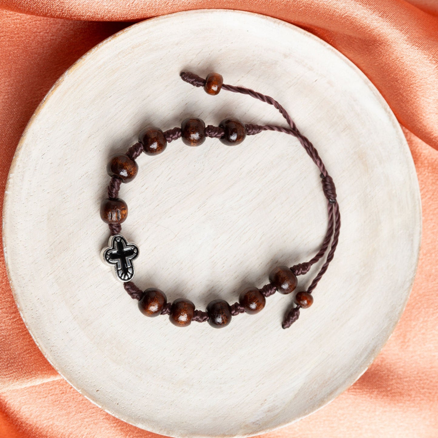 A beaded bracelet with dark wooden beads and a silver cross charm, displayed on a copper fabric background with decorative elements.