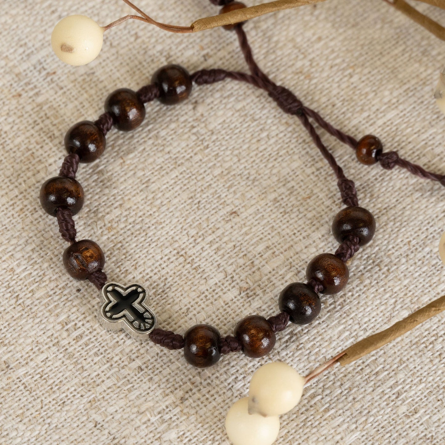 A beaded bracelet with dark wooden beads and a silver cross charm, displayed on a beige background with decorative elements.