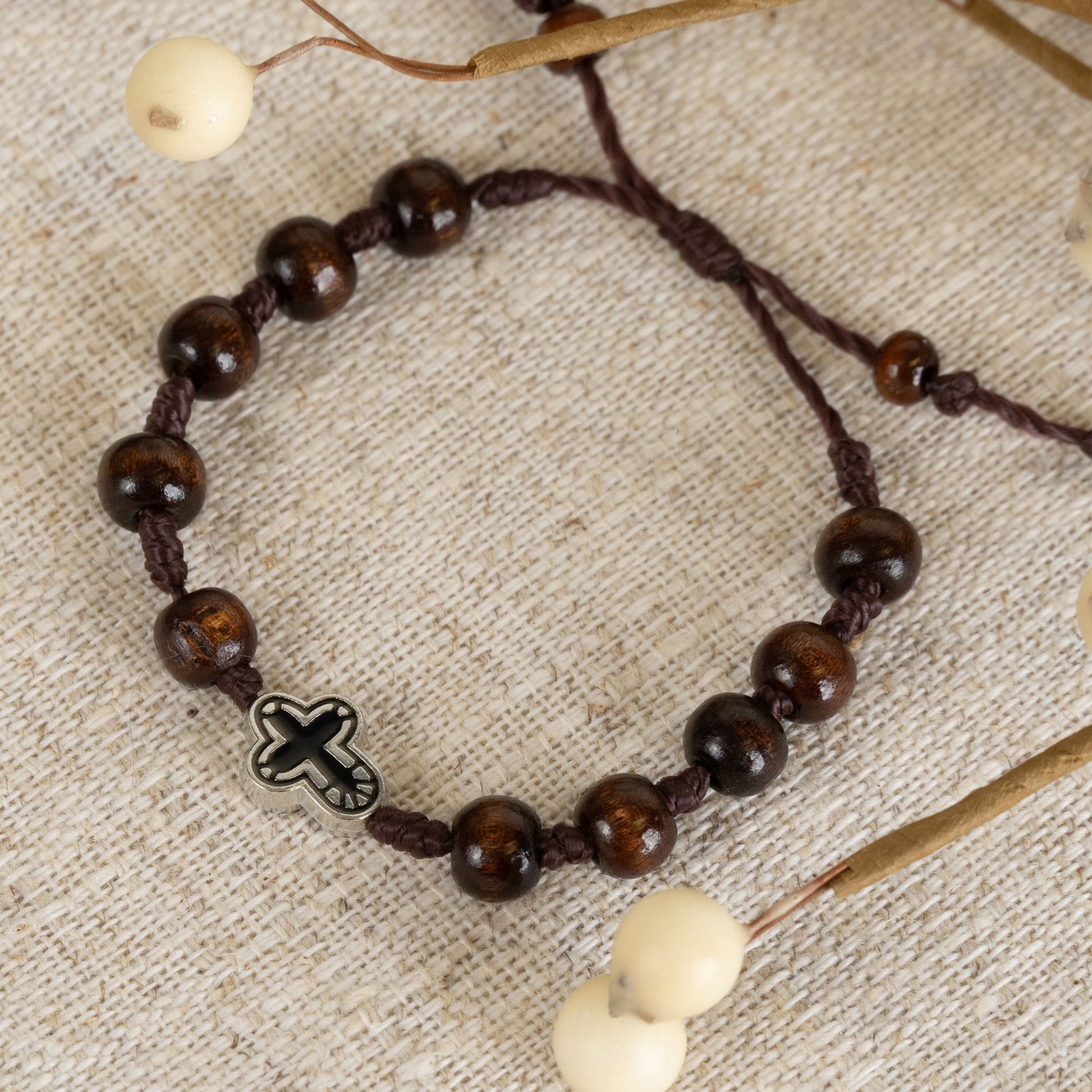 A beaded bracelet with dark wooden beads and a silver cross charm, displayed on a beige background with decorative elements.