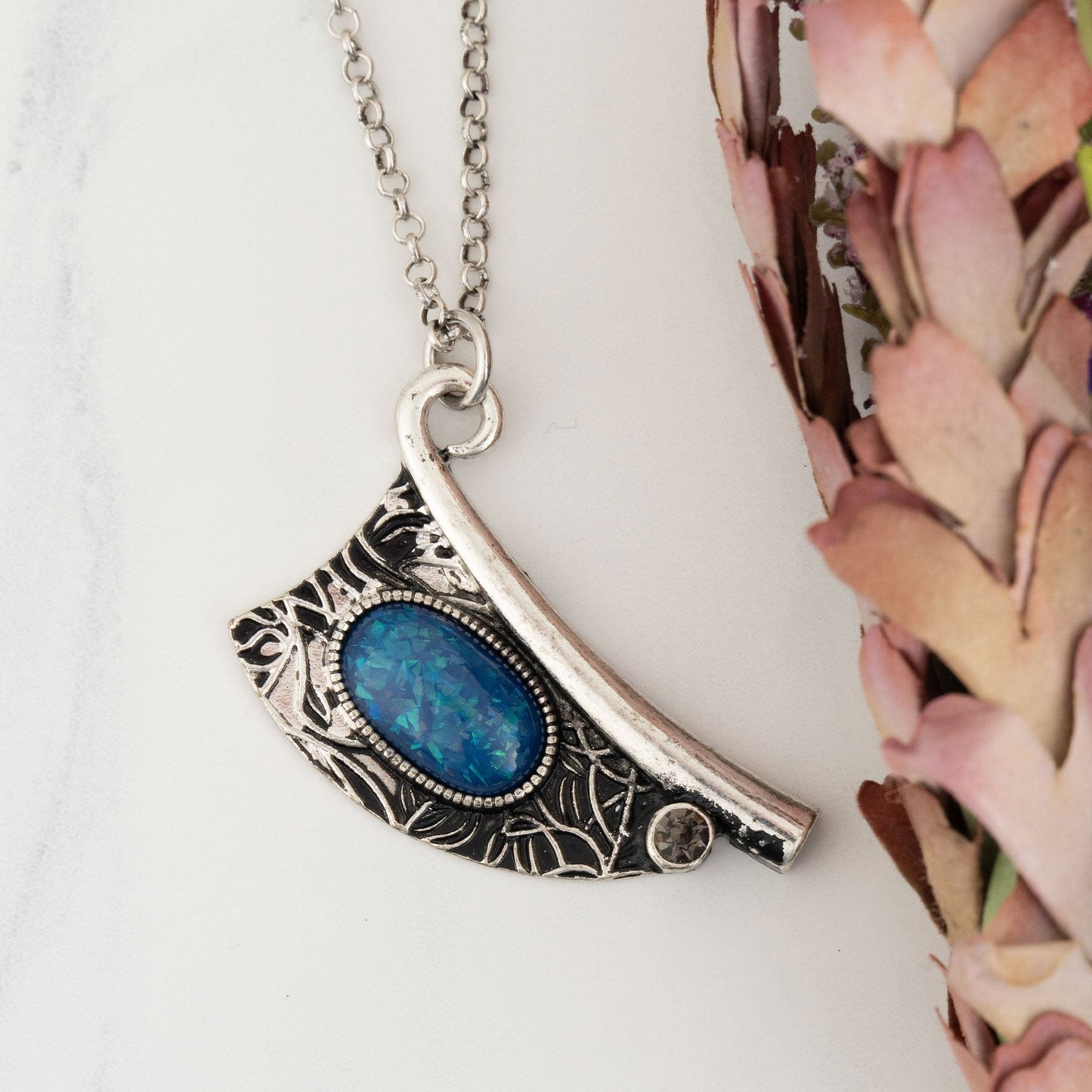 A silver pendant necklace with an intricate engraved design and a vibrant blue bead centered on the pendant, displayed against a floral background.