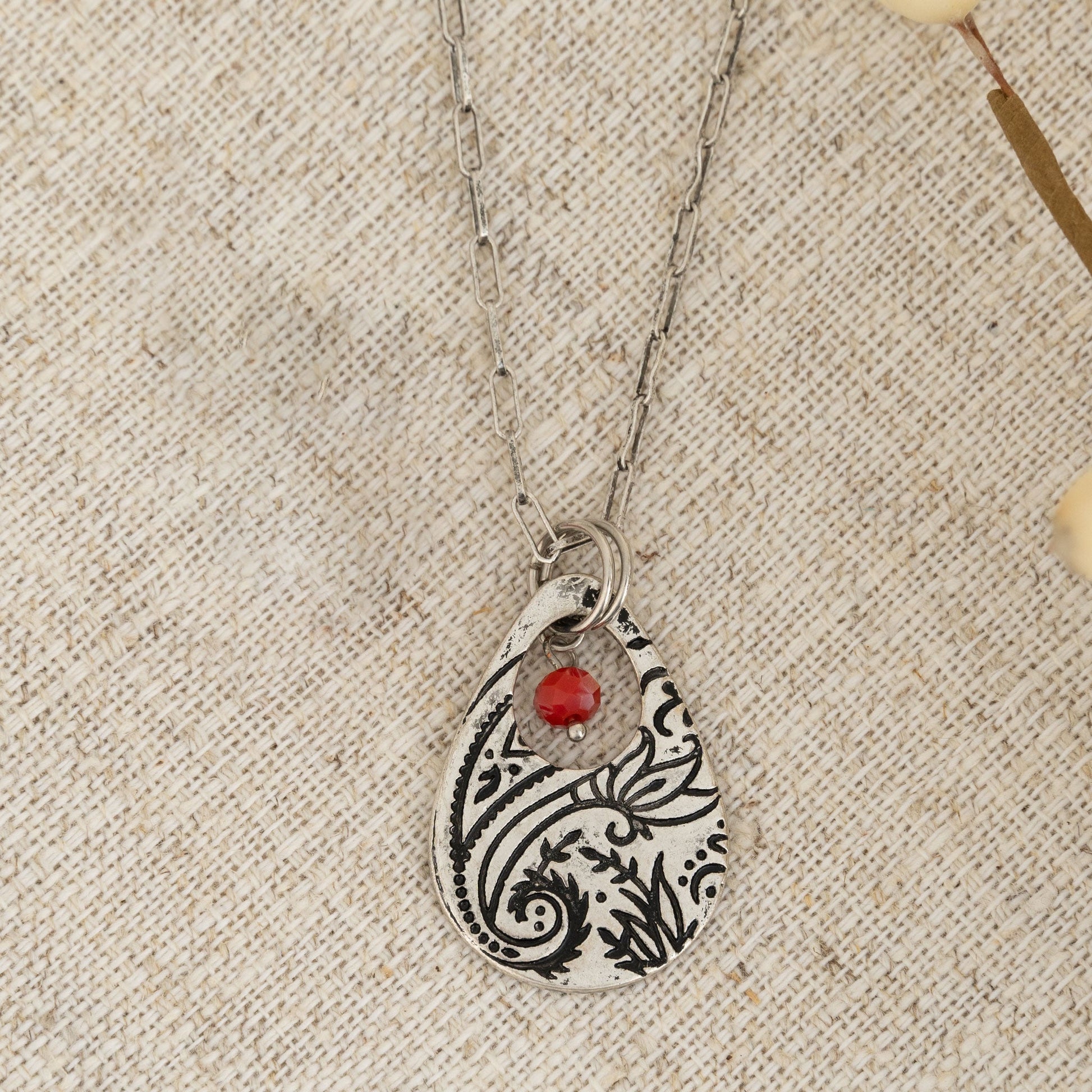 A silver chain pendant necklace with a teardrop-shaped paisley design pendant and a red bead on a textured background.