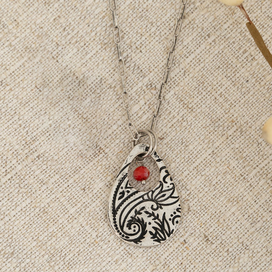 A silver chain pendant necklace with a teardrop-shaped paisley design pendant and a red bead on a textured background.