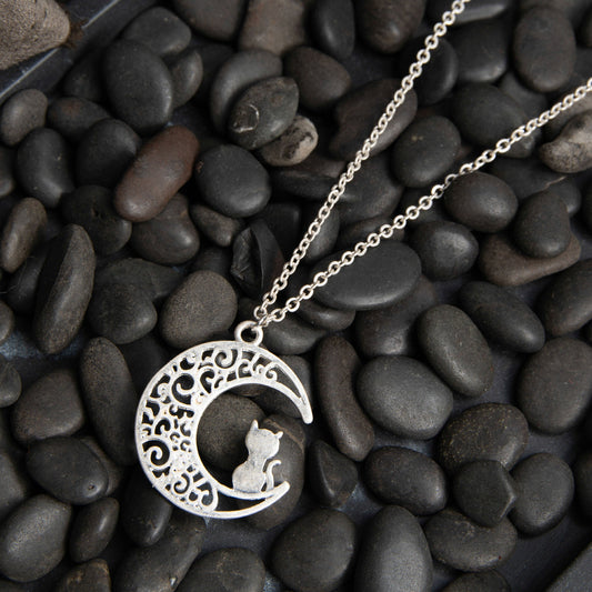 A silver necklace with a filigree crescent moon and cat silhouette pendant lying on a bed of pebbles.
