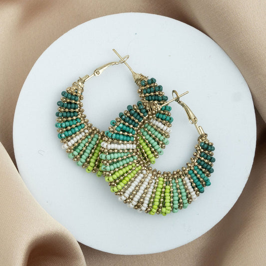 A pair of seed bead hoop earrings with green and gold colors, displayed on a white plate.