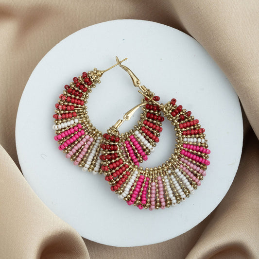 A pair of gold-colored hoop earrings with red and white seed beads wrapped around them, displayed on a white plate with a satin background.