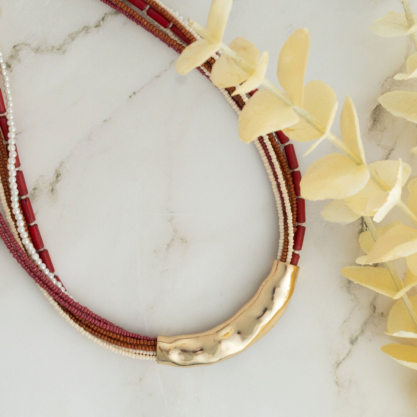 A seed bead necklace in red and gold colors with hammered metal detailing, displayed on a marble surface.