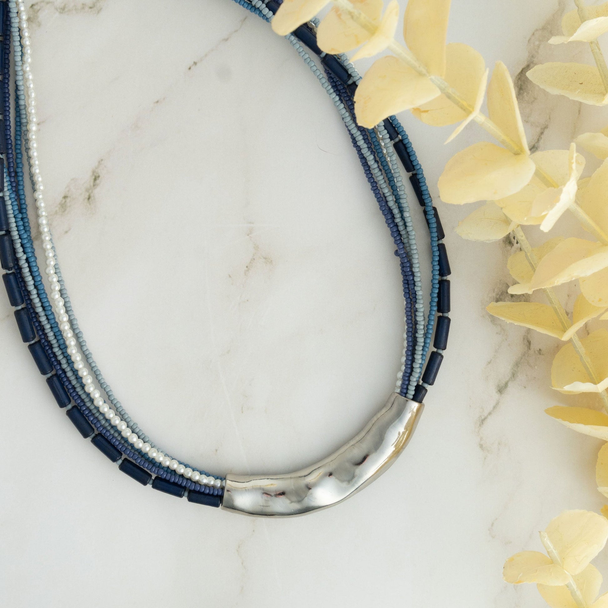 A blue seed bead necklace with silver hammered detailing, displayed on a marble surface with yellow flowers in the background.