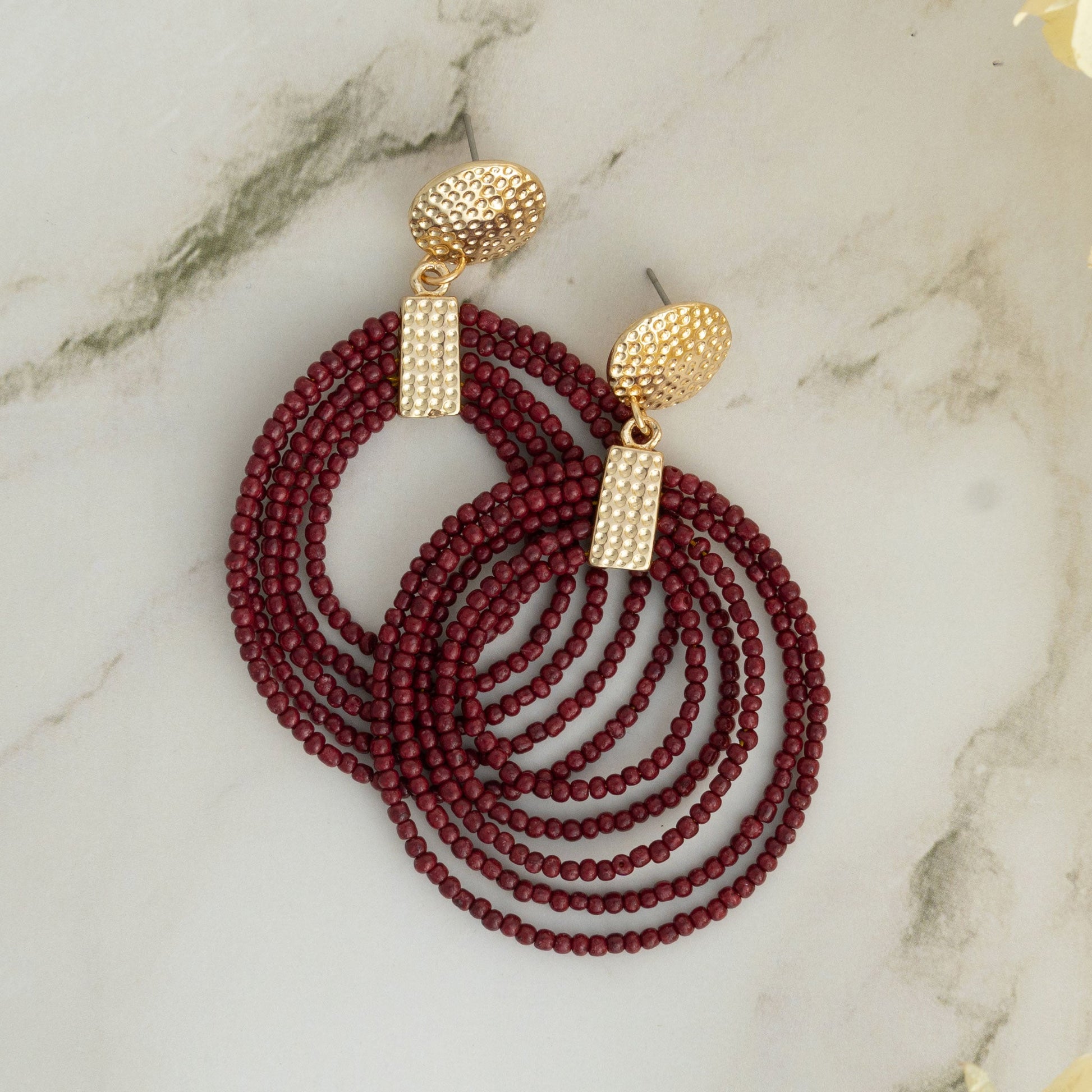 A pair of gold-colored hoop earrings featuring red seed bead detailing, displayed on a marble surface.