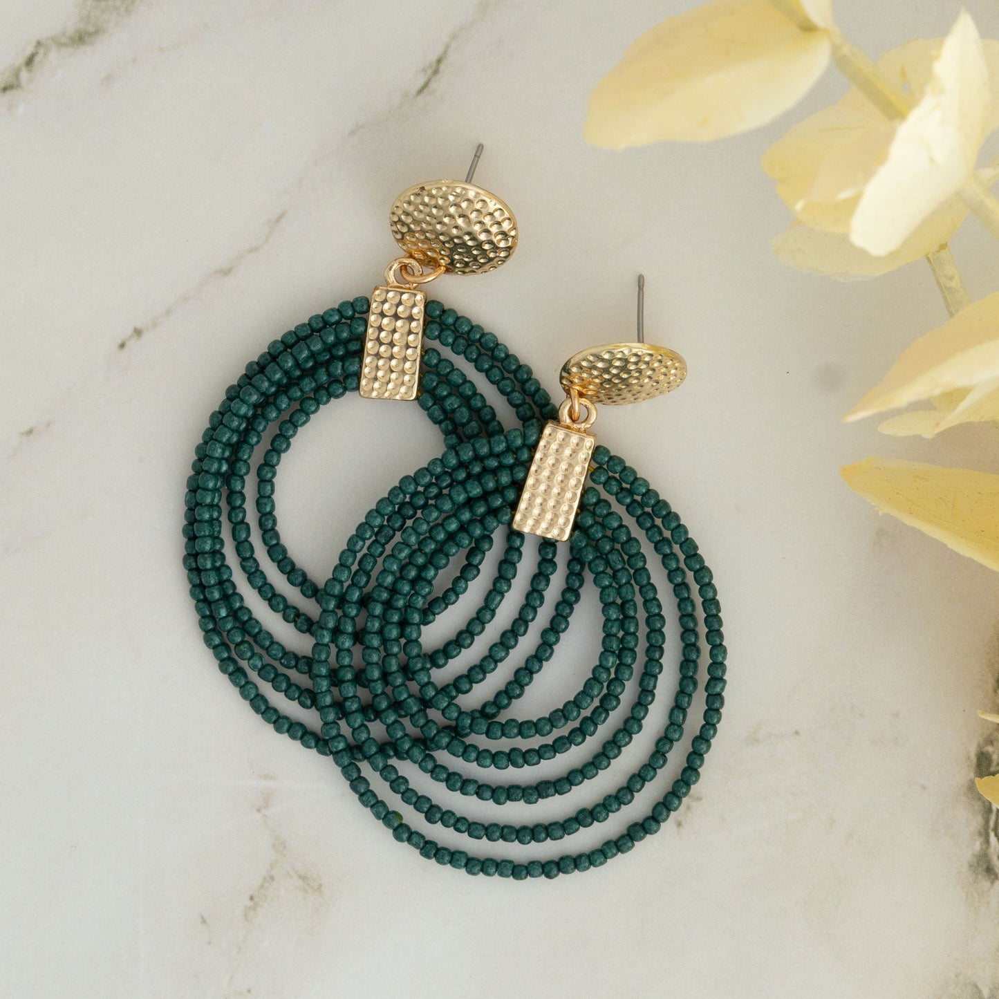 A pair of hoop earrings made of green seed beads with gold detailing at the top, displayed on a marble surface.