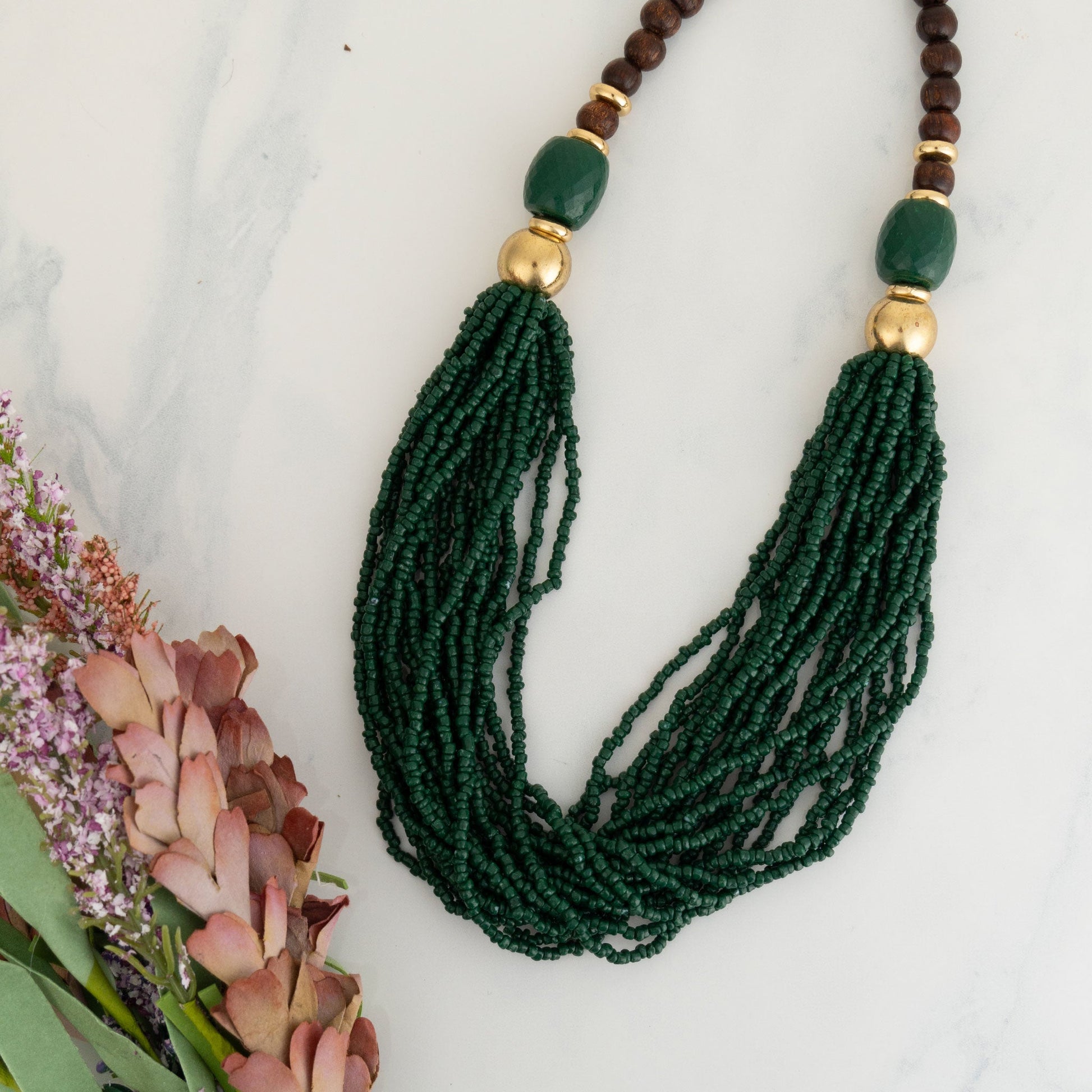 A dark green seed bead necklace with gold-tone accents displayed on a marble surface next to some flowers.