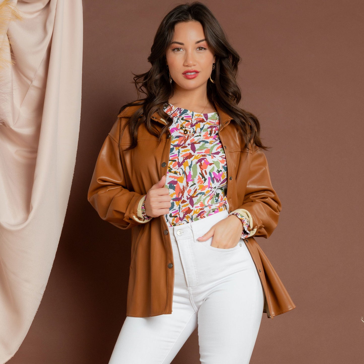 A woman wearing a multicolored balloon sleeve blouse with a round neckline, paired with a faux leather jacket and white jeans
