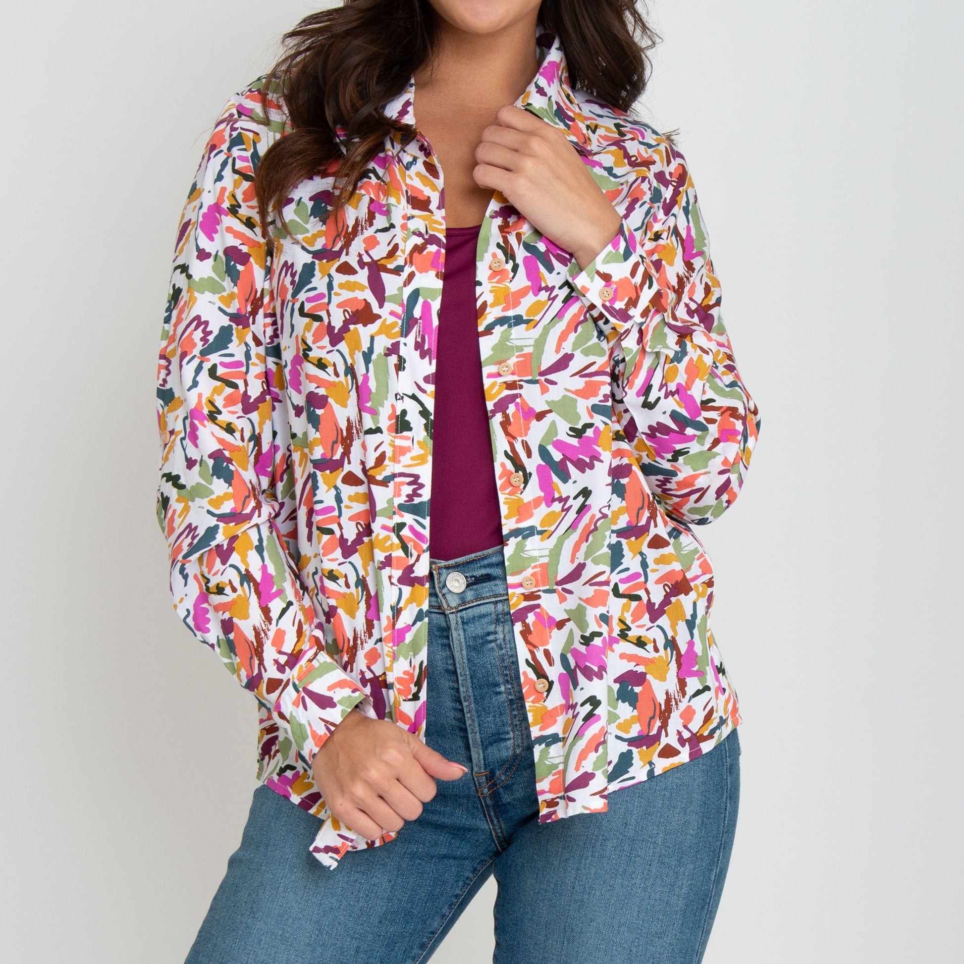 A woman wearing a multicolor button-up shirt with long sleeves, paired with blue jeans and a purple top, standing against a neutral background.