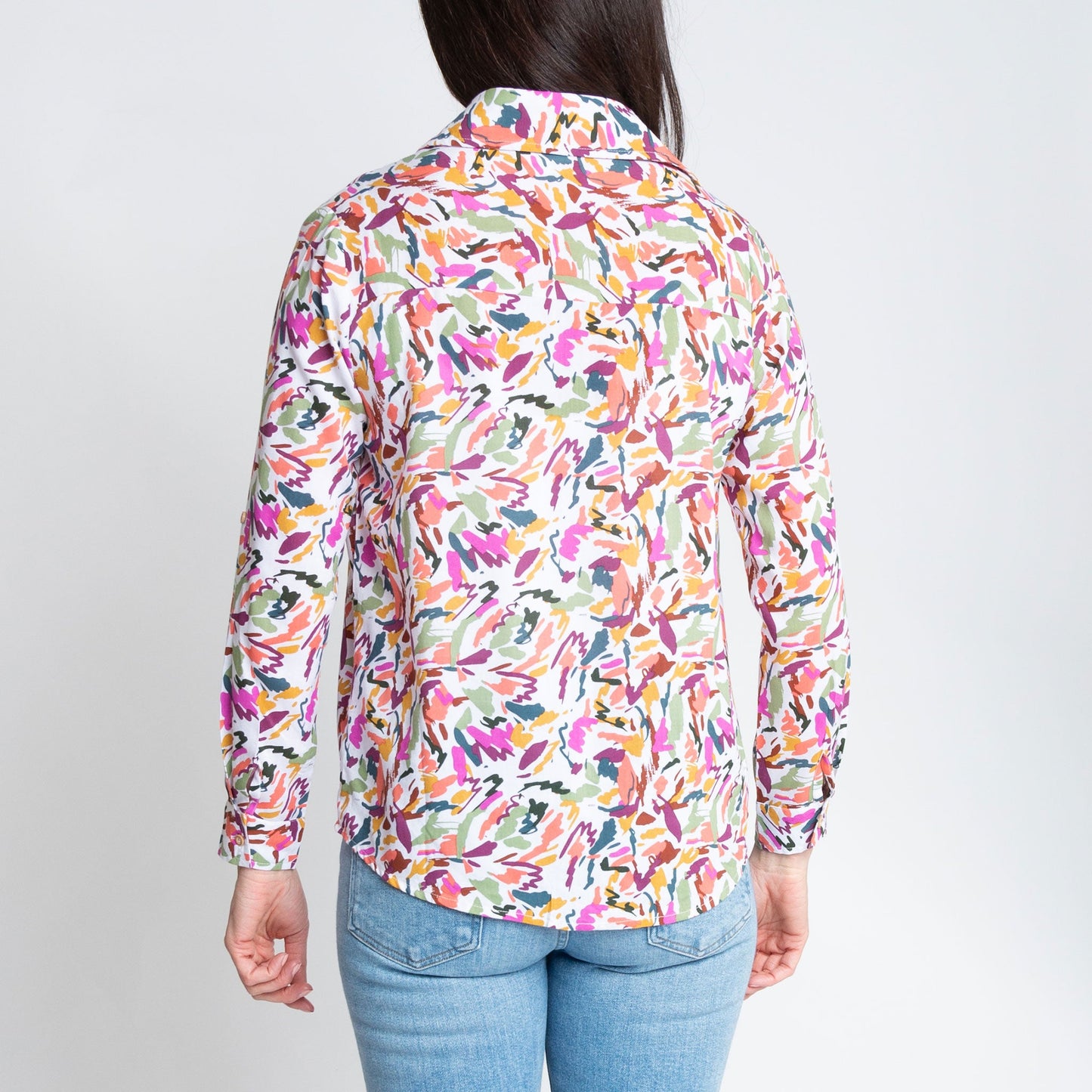 A woman wearing a multicolor button-up shirt with long sleeves, paired with blue jeans, standing against a neutral background.
