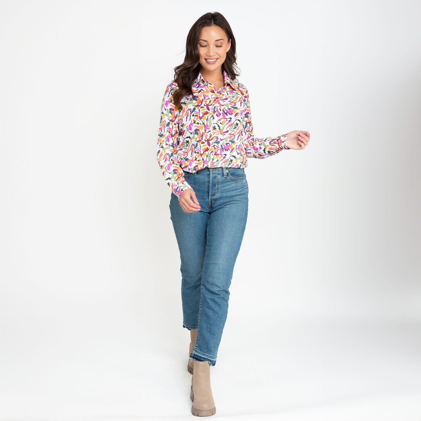 A woman wearing a multicolor button-up shirt with long sleeves, paired with blue jeans, standing against a neutral background.