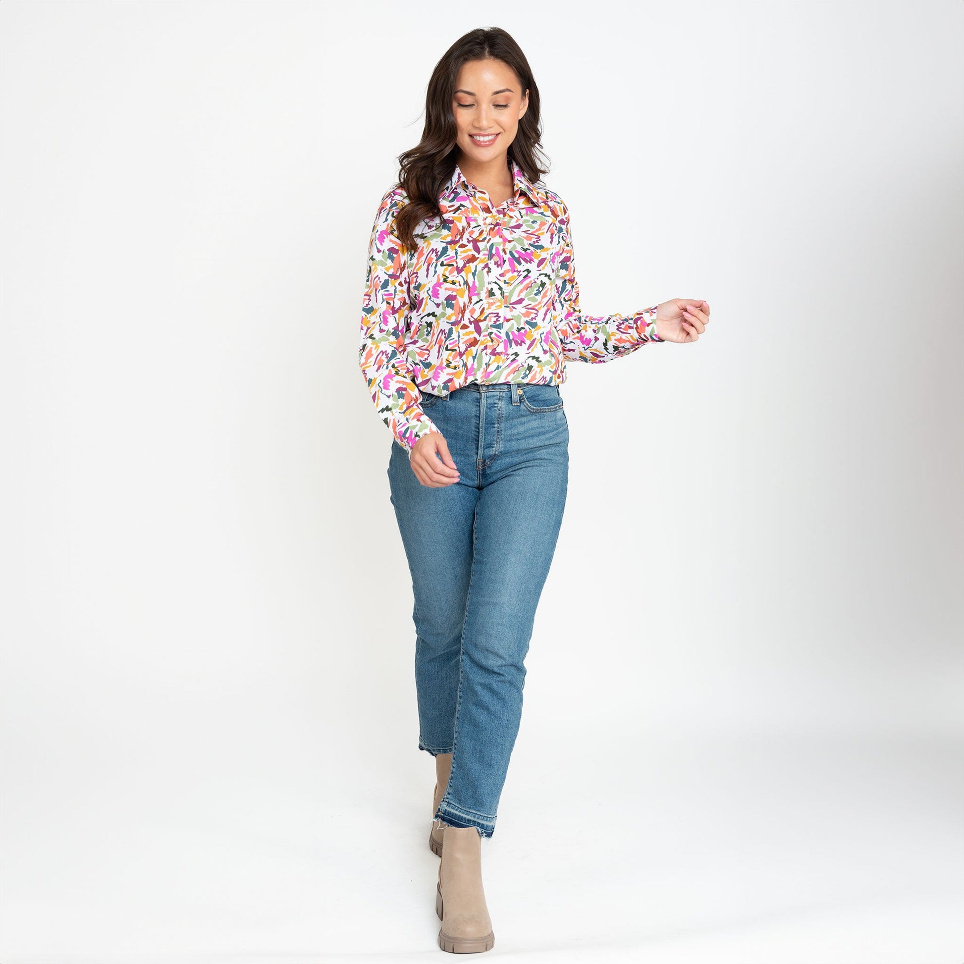 A woman wearing a multicolor button-up shirt with long sleeves, paired with blue jeans, standing against a neutral background.