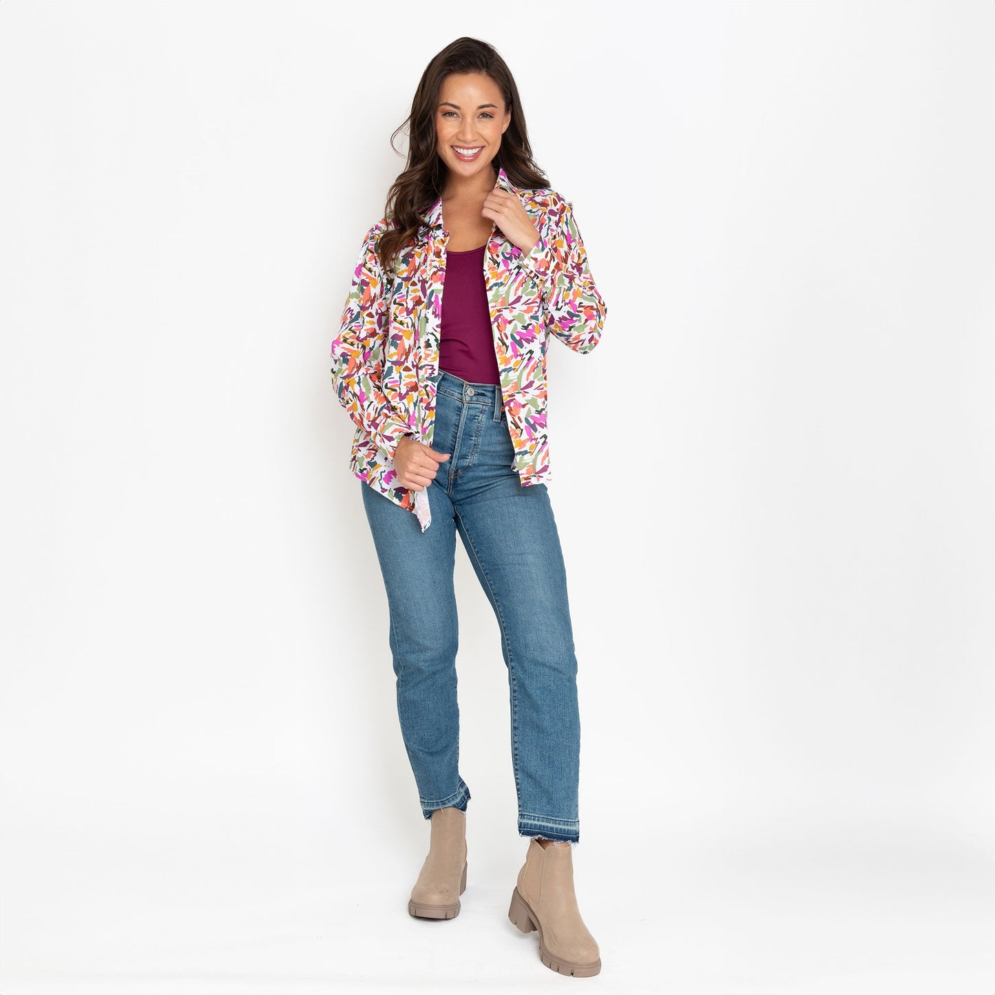 A woman wearing a multicolor button-up shirt with long sleeves, paired with blue jeans, standing against a neutral background.