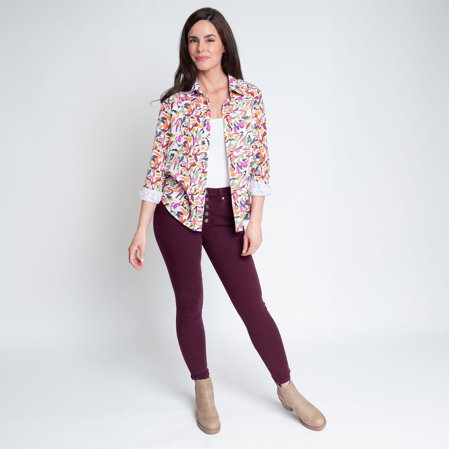 A woman wearing a multicolor button-up shirt with long sleeves, paired with purple jeans, standing against a neutral background.