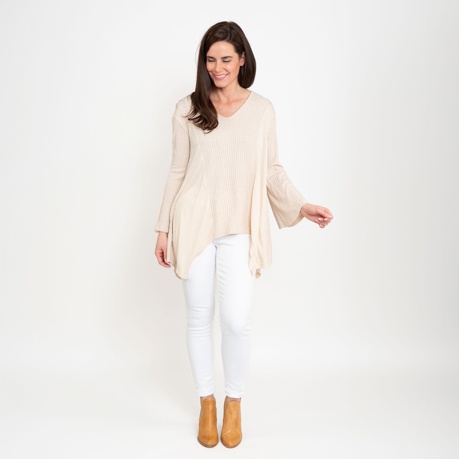 A woman wearing a beige Cassia Bell Sleeve Top with a V-neck and long bell sleeves, paired with white jeans.