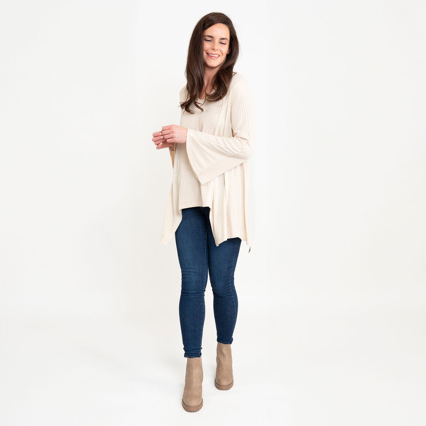 A woman wearing a beige Cassia Bell Sleeve Top with a V-neck and long bell sleeves, paired with blue jeans.