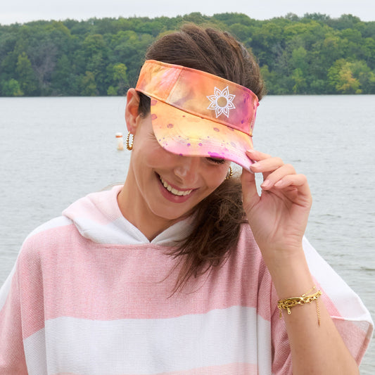 A woman wearing a pink and orange tie dye visor with a mandala print icon in front of a beach.