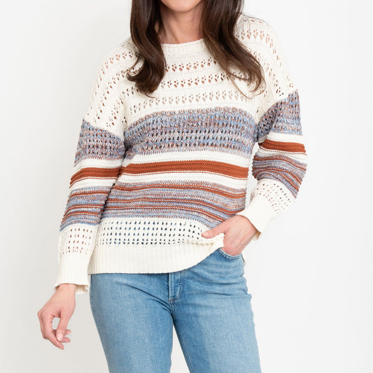 A woman wearing a color block crochet knit sweater with a boatneck neckline paired with jeans.